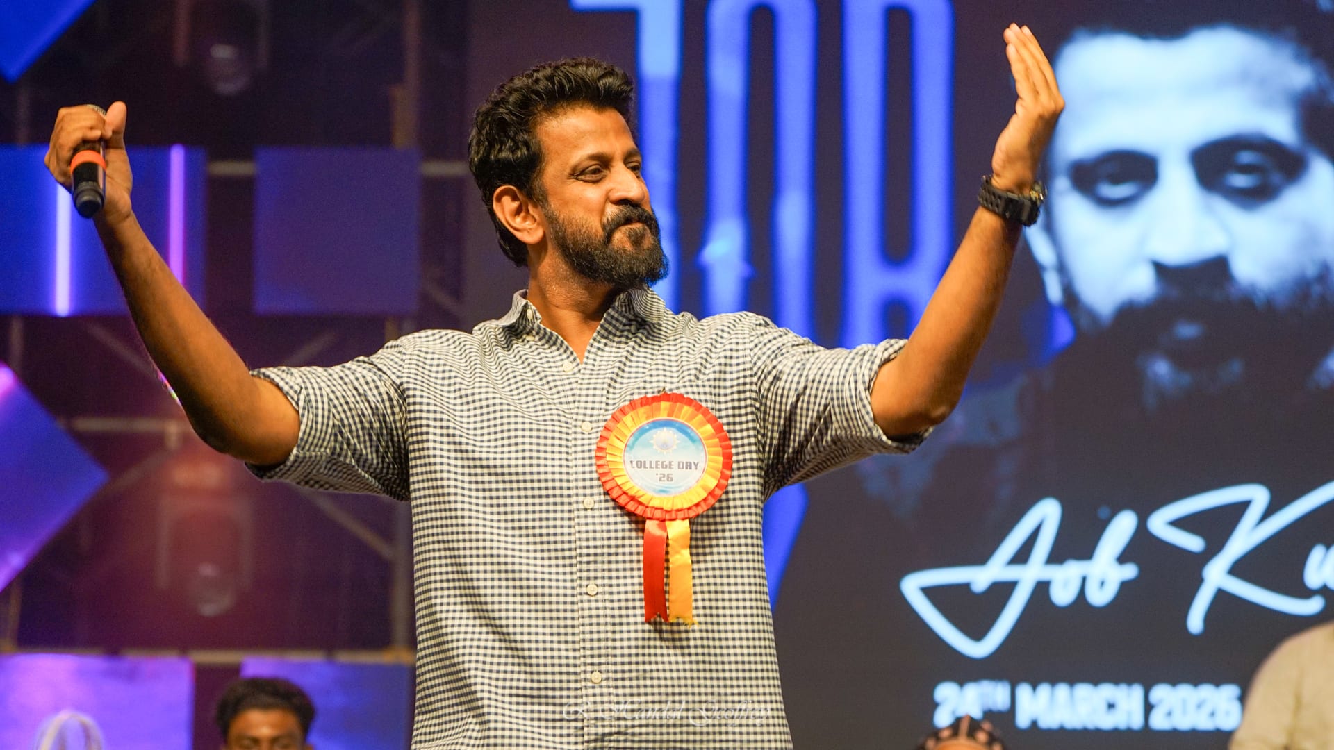 Job Kurian on stage at College Day, arms raised, microphone in hand